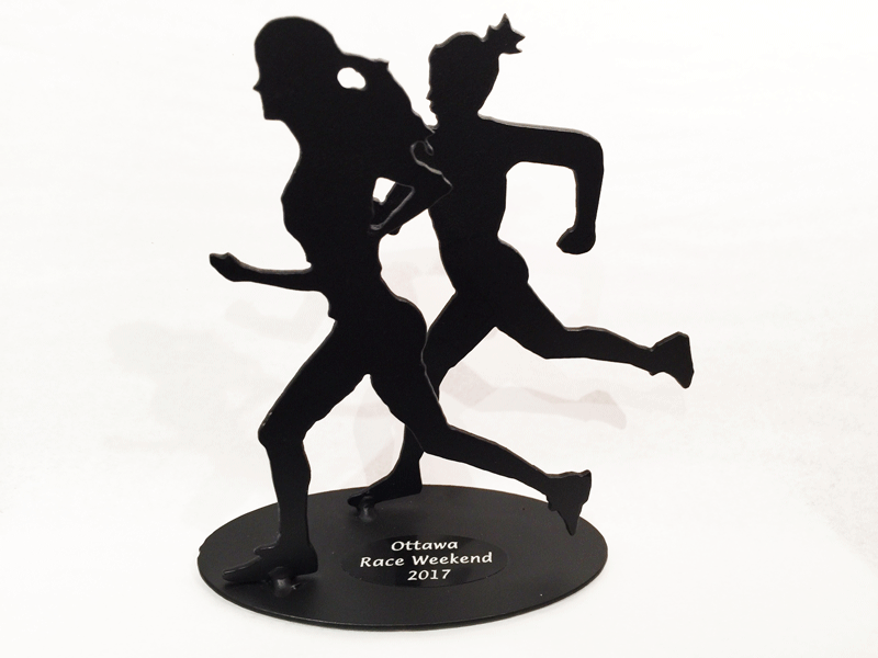 A metal sculpture of two female runners. The sculpture is painted matte black. A small black metal plate has been attached to the base of the sculpture. “Ottawa Race Weekend 2017” is engraved in bright silver on the plate.