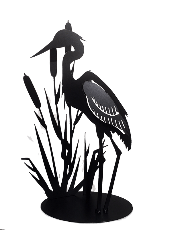 This metal sculpture shows the matte black silhouette of a heron standing with cattails. The wing of the heron has been partially punched out of the metal, and bent to render the bird in 3D. Individual wing feathers are visible. The cattails are cut very delicately and in lifelike shape and proportion to the heron. The piece sits on an oval base.