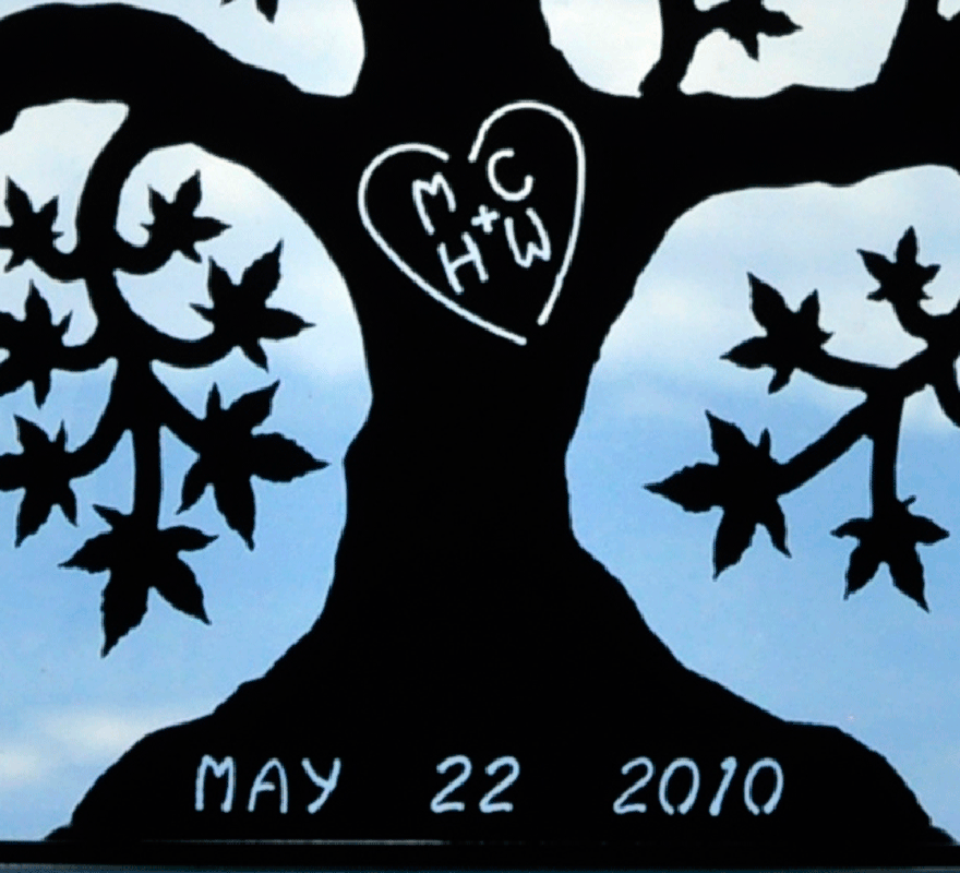 A close up of a matte black tree of love. The initials “MC + HW” are carved into the trunk, enclosed by a heart. The date “May 22, 2010” is carved into the base. This photo highlights the contrast between the matte black sculpture and the bright light passing through the carving.
