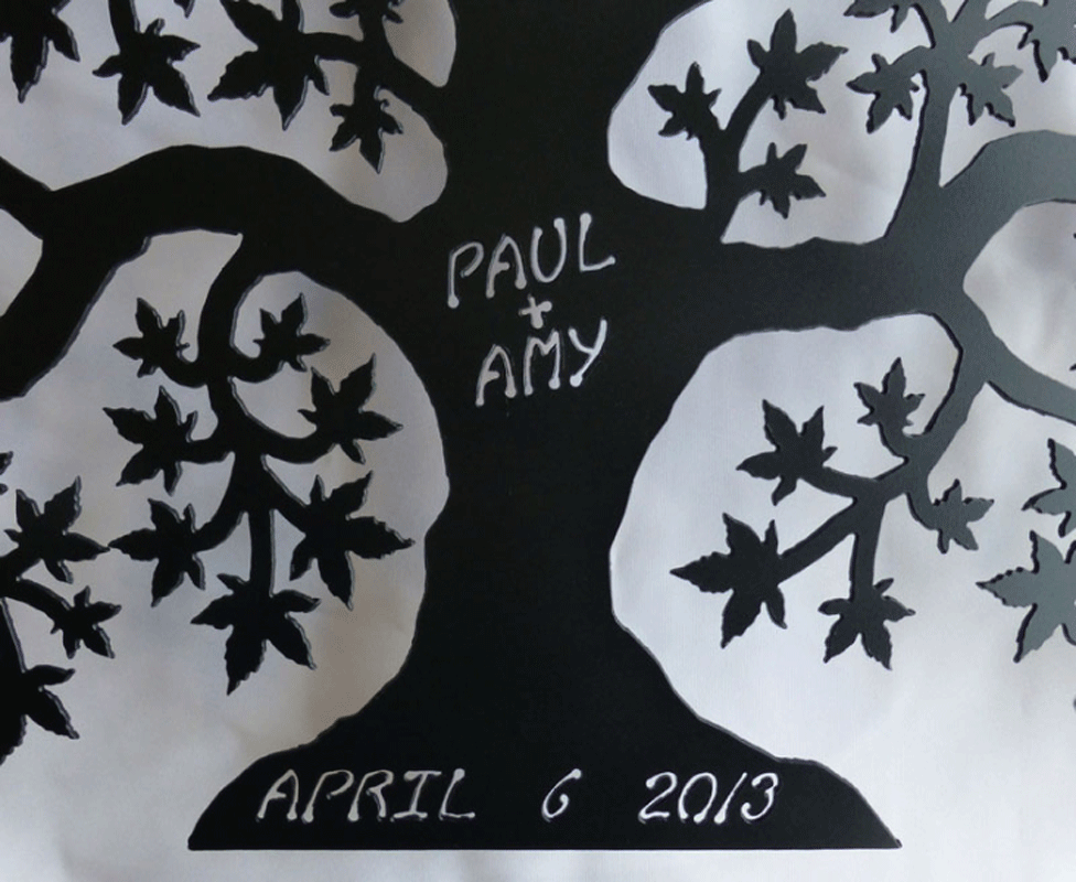 A close up of a matte black tree of love. The names “Paul + Amy” are carved into the trunk. The date “April 6 2013” is carved into the base of the tree. 