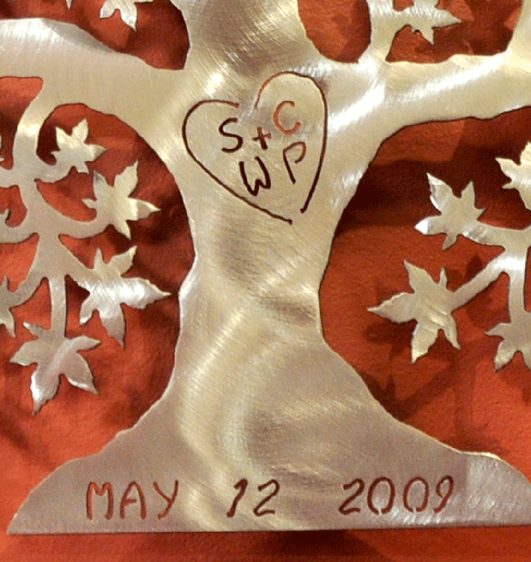 A close up of a brushed metal tree of love on a brown wall. The initials “SC + WP” are carved into the trunk, enclosed by a heart. The date “May 12, 2009” is carved into the base. Although more subtle, the brushed metal tree still contrasts significantly with the darker background, allowing good legibility of the text.