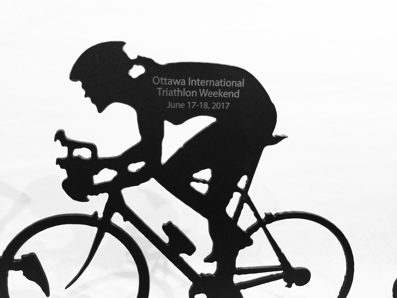 A close up of the engraving on the female triathlete sculpture. Direct engraving on her back reads "Ottawa International Triathlon Weekend June 17-18, 2017" in grey contrasting against the black paint of the figure.