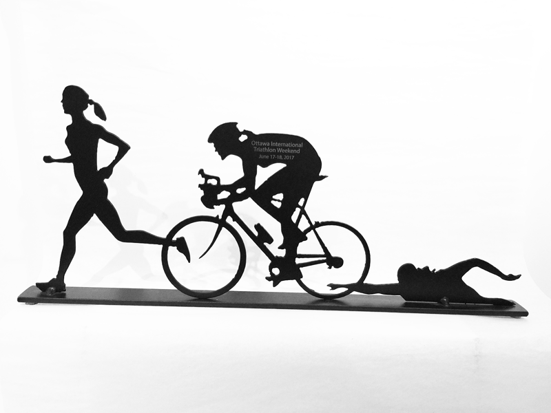 A metal sculpture of three female athletes, each performing one challenge of a triathlon. The sculpture is painted matte black. Across the middle figure’s back is engraved “Ottawa International Triathlon Weekend June 17-18, 2017”. The direct engraving allows the grey metal underlying  to show against the black paint.