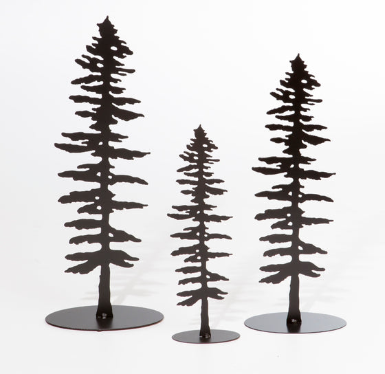 Three metal sculptures each show the matte black silhouette of a Sitka pine tree.  The designs of each tree are identical, but each sculpture is a different size, with the largest on the left and the smallest in the center. The Sitka tree is tall but slim. Its short, broad branches are about the same length along the whole tree, except at the top where they form a point. The sculpture sits on an oval base.