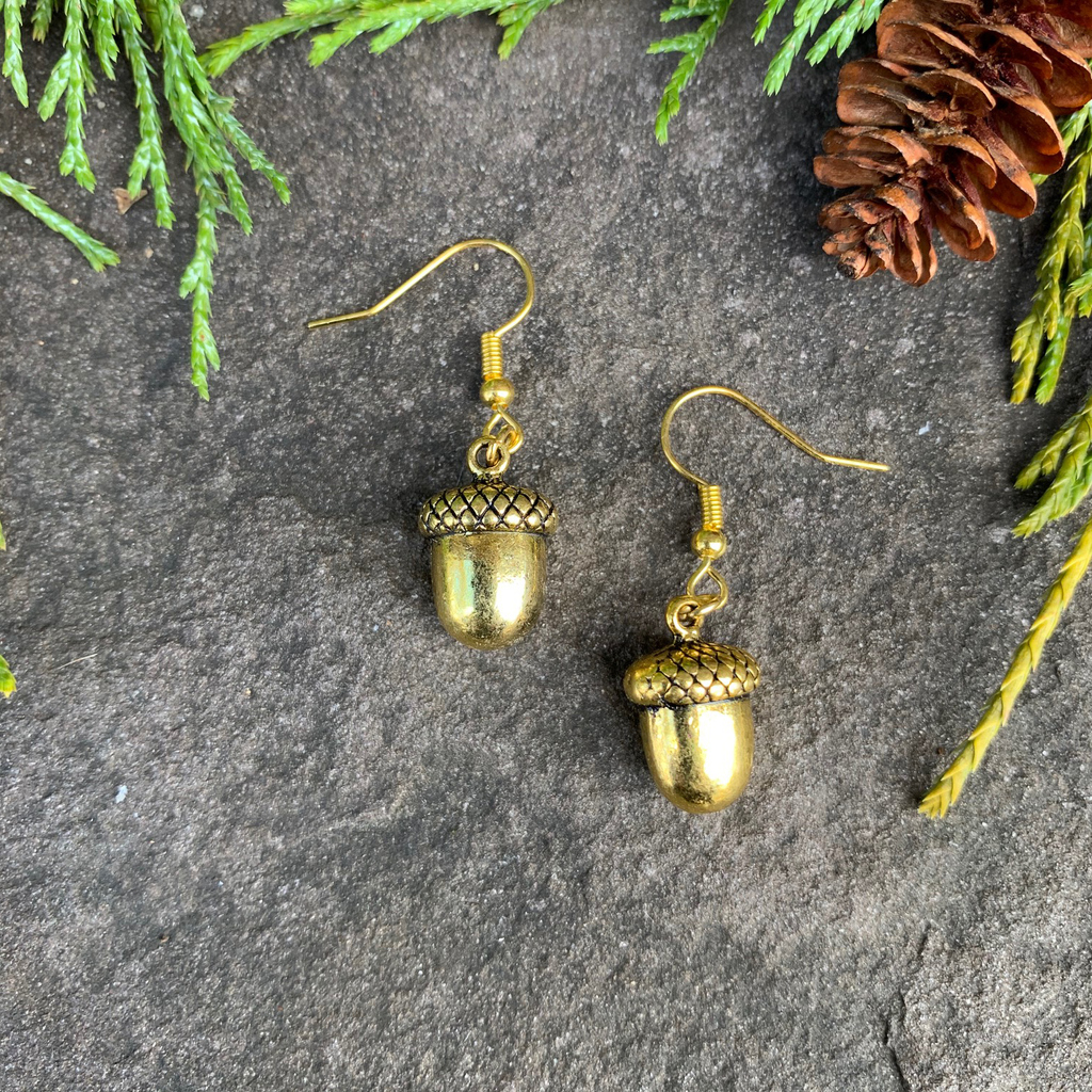 Two small gold acorn charms sit on a stone background. A gold earring hook is attached to the top of each acorn. 