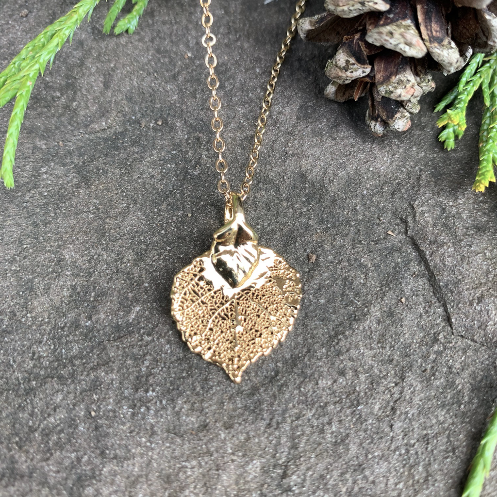 A small gold coated aspen leaf on a gold chain sits on a stone background. The gold has a bright finish. At the top of the picture are decorative evergreen leaves and a pine cone.
