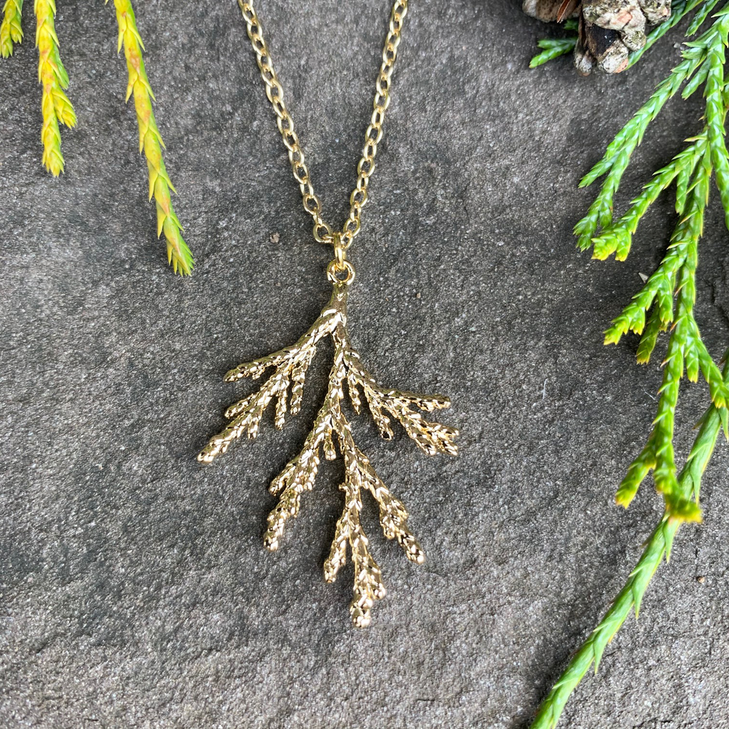 A small gold coated piece of juniper on a gold chain sits on a stone background. The gold has a bright finish. At the top of the picture are decorative evergreen leaves and a pine cone.
