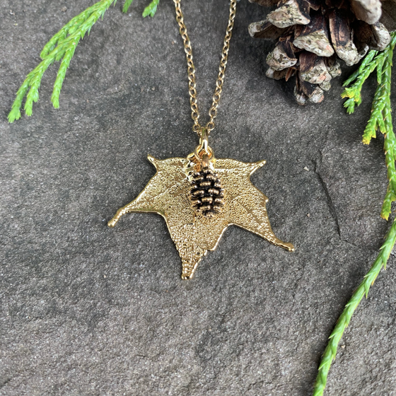 A small gold coated maple leaf on a gold chain sits on a stone background. Attached to the maple leaf is a tiny gold pinecone. The gold has a bright finish. At the top of the picture are decorative evergreen leaves and a pine cone.