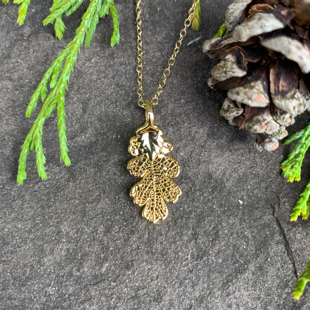 A small gold coated oak leaf on a gold chain sits on a stone background. The gold has a bright finish. Around the picture are decorative evergreen leaves and a pine cone.