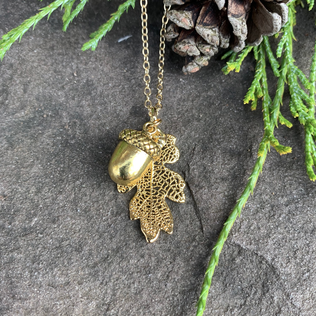 A small gold coated oak leaf on a gold chain sits on a stone background.  Also attached to the chain is a small gold acorn charm. The gold has a bright finish. Around the picture are decorative evergreen leaves and a pine cone.
