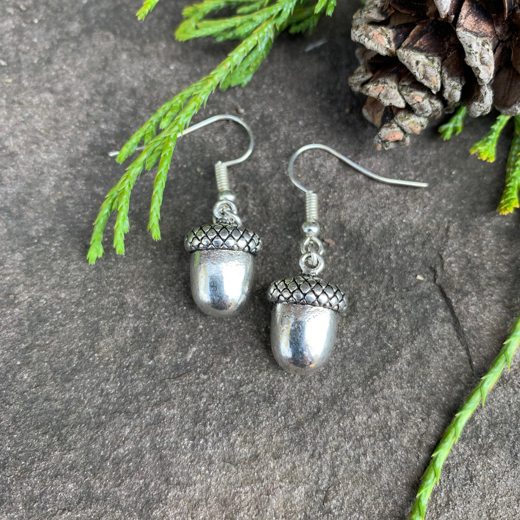 Two small silver acorn charms sit on a stone background. A silver earring hook is attached to the top of each acorn. 