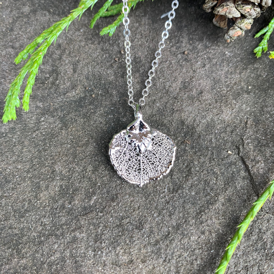 A small silver coated aspen leaf on a silver chain sits on a stone background. The silver has a bright finish. At the top of the picture are decorative evergreen leaves and a pine cone.