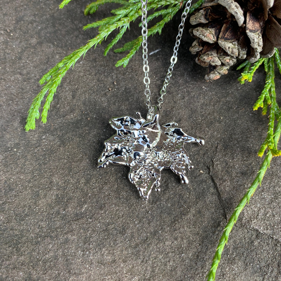A small silver coated maple leaf on a silver chain sits on a stone background. The silver has a bright finish. At the top of the picture are decorative evergreen leaves and a pine cone.