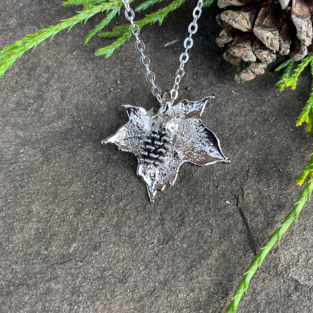 A small silver coated maple leaf on a silver chain sits on a stone background. Attached to the maple leaf is a tiny silver pinecone. The silver has a bright finish. At the top of the picture are decorative evergreen leaves and a pine cone.