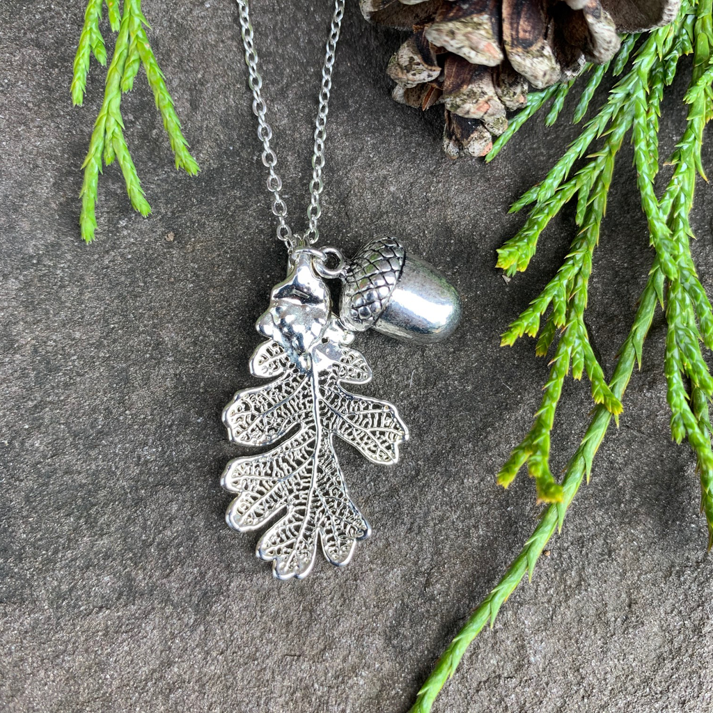 A small silver coated oak leaf on a silver chain sits on a stone background.  Also attached to the chain is a small silver acorn charm. The silver has a bright finish. Around the picture are decorative evergreen leaves and a pine cone.