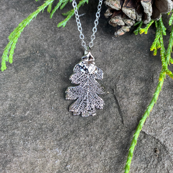 A small silver coated oak leaf on a silver chain sits on a stone background. The silver has a bright finish. Around the picture are decorative evergreen leaves and a pine cone.