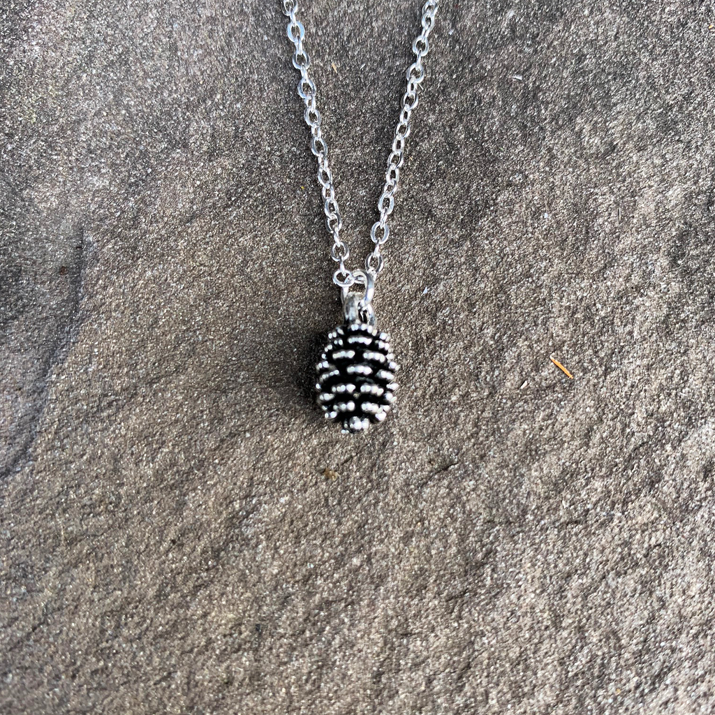 Sliver cable chain with small oval shaped pinecone pendant. Pinecone has shiny finish and several grooves that give a realistic appearance. Necklace sits on stone background.