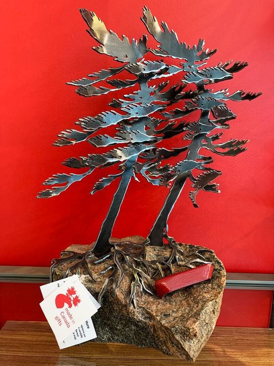 Steel windswept pine trees fitted onto a piece Canadian stone. A red steel canoe sits on the rock next to the pine tree roots.