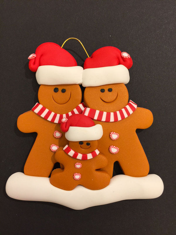 Two smiling gingerbread men stand side by side wearing a red Santa hat, red and white mints as buttons, and a red and white striped scarf. Standing between them is a smaller gingerbread child.