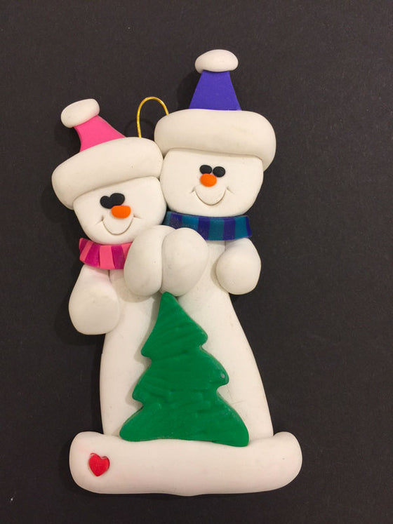 Two smiling snowmen hold hands. The one on the left wears a pink hat and scarf and the one on the right wears a blue hat and scarf. A small Christmas tree sits in front of them.