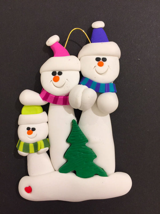 Two smiling snowmen hold hands. The one on the left wears a pink hat and scarf and the one on the right wears a blue hat and scarf. A small Christmas tree sits in front of them.  The pink snowman places its other hand on the head of a smaller snowman dressed in green.
