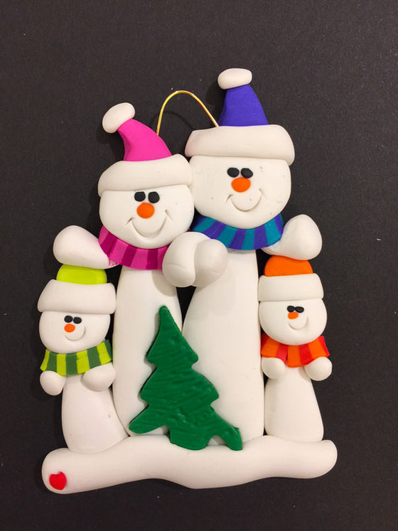 Two smiling snowmen hold hands. The one on the left wears a pink hat and scarf and the one on the right wears a blue hat and scarf. A small Christmas tree sits in front of them.  The pink snowman places its other hand on the head of a smaller snowman dressed in green. The blue snowman does the same with a small orange snowman.