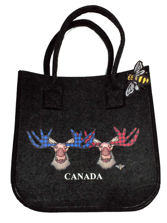 This small dark grey felt bag has two overhead handles and features an art print of two moose. The left moose’s antlers are coloured with blue buffalo check, while the right moose has red buffalo check. The moose’s ear, nose, face and neck are all coloured with different shades of brown. Underneath the moose the word Canada has been written in white text. At the bottom right of the picture is the artists mark—a small picture of a bee.