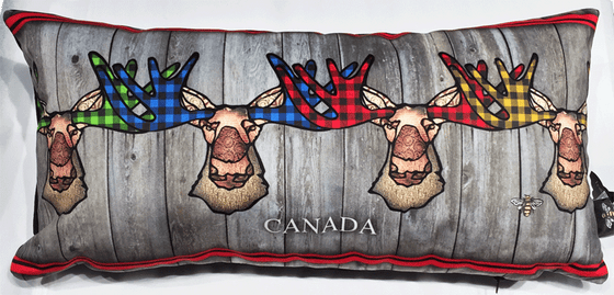 This large pillow features four funky moose. The moose’s ear, nose, face and neck are all coloured with different shades of brown. Each moose’s antlers are coloured with either green, blue, red, or yellow buffalo check. A decorative red stripe runs along the top and bottom of the pillow. Underneath the moose the word Canada has been written in white text. At the bottom right of the pillow is the artists mark—a small picture of a bee.
