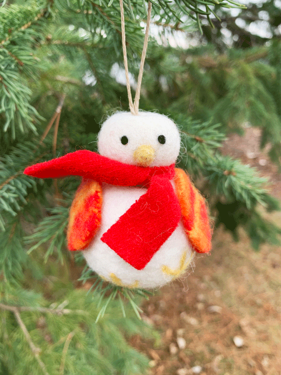 A felted snowbird. It has two black eyes and a yellow cone beak, and two embroidered feet on its round body. Its wings are mottled red and yellow and it wears a red scarf.