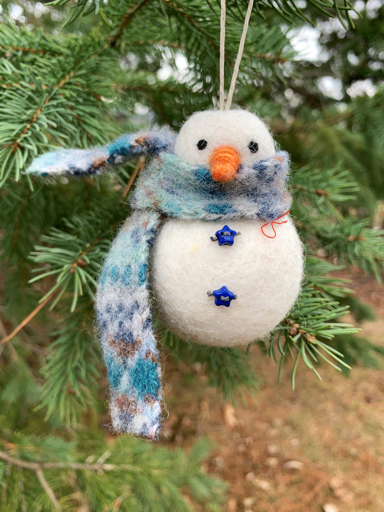 A felted snowman with a head and body segment. The head has two small black eyes and an orange carrot nose. It has a mottled blue scarf and two blue starshaped buttons on its belly.
