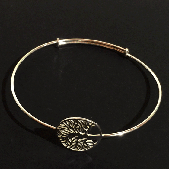 A sterling silver tree charm attached to a thin silver bracelet. The tree is contained within a hoop. The tree’s numerous thin branches radiate outward and fuse with the outer hoop. The charm is attached to the bracelet along the back of the tree trunk. The bracelet may expand to fit a larger wrist, if necessary.