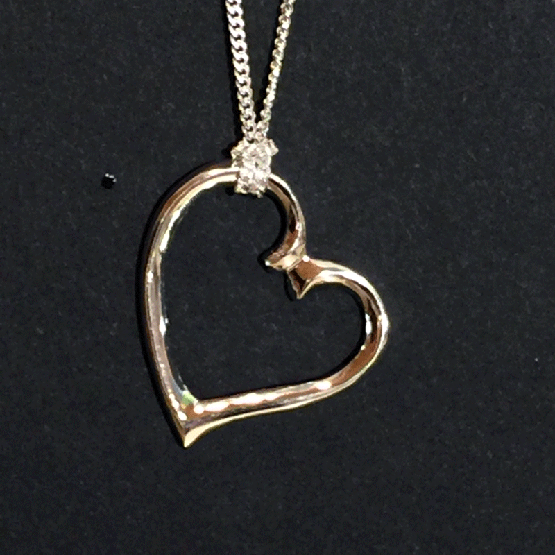 A sterling silver heart charm hangs from a silver chain on a black background. The chain attaches to the top left of the heart. The heart is asymmetrical and appears to curl towards the right.