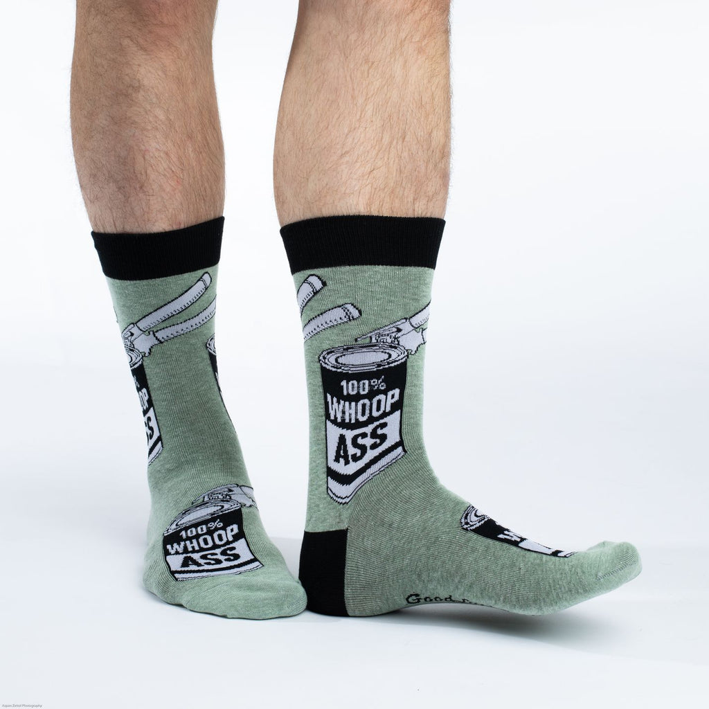 These fun socks feature a can with a label that says “ 100% WHOOP ASS”, and can opener on the can. The images are on a speckled olive-green background with a black heel and rim. Spandex added to the 85% cotton blend gives the socks the perfect amount of stretch to hug your feet.