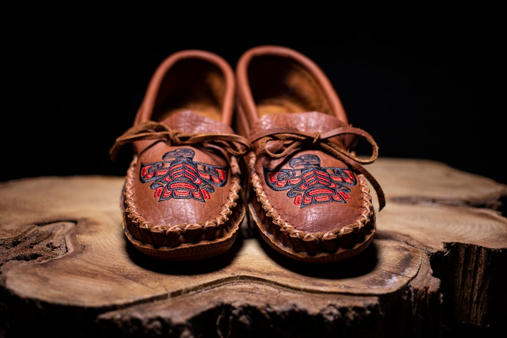 Dark brown moccasins with embroidered Haida Eagle on the vamp with a lace to adjust it. Moccasins on sitting on a tree stump.