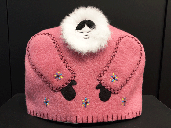 This tea cozy is in the shape of a smiling person wears a pink parka with white fur around their face. Darker pink stitching outlines the sleeves. Two brown mittens poke out from the sleeves, and blue and yellow stars are stitched along the bottom and on the sleeves. 