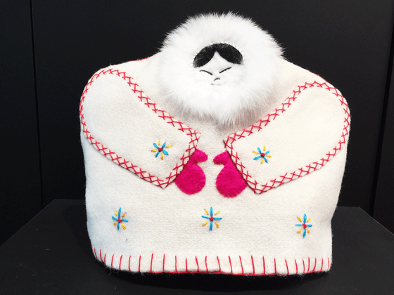 This tea cozy is in the shape of a smiling person wearing a white parka with white fur around their face. Red stitching outlines the sleeves, and pink mittens poke out from the end of them. blue and yellow stars are stitched onto the parka, three along the bottom, and one on each sleeve.