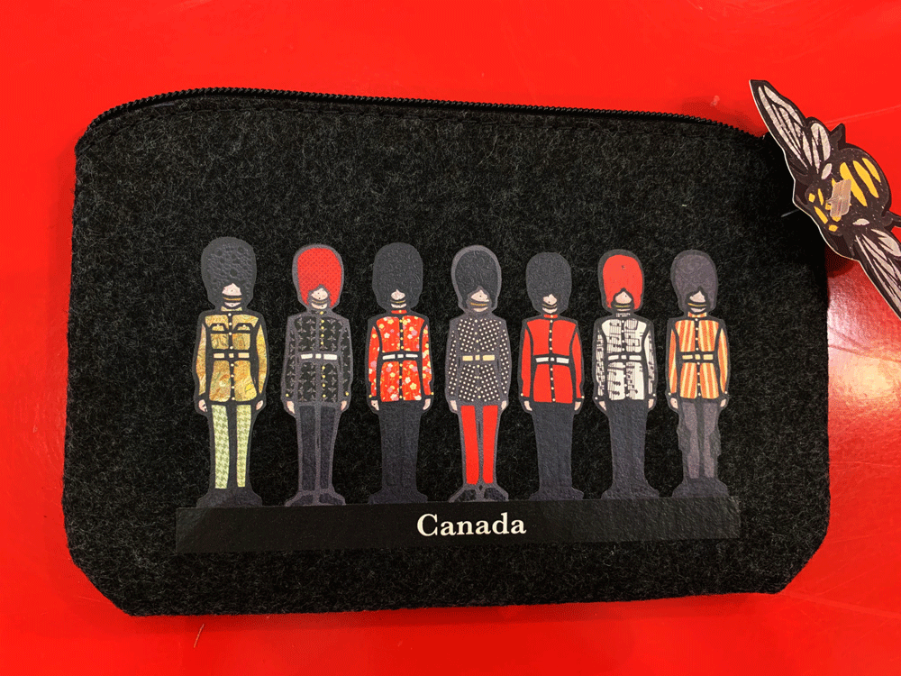 This small dark grey pouch has an overhead zipper and features an art print of seven soldiers standing at attention. They are al wearing long pants, belted jackets, and tall beefeater hats. Their jackets each have a unique colourful patterned print on them. At the bottom of the picture the word Canada is written in white text. 