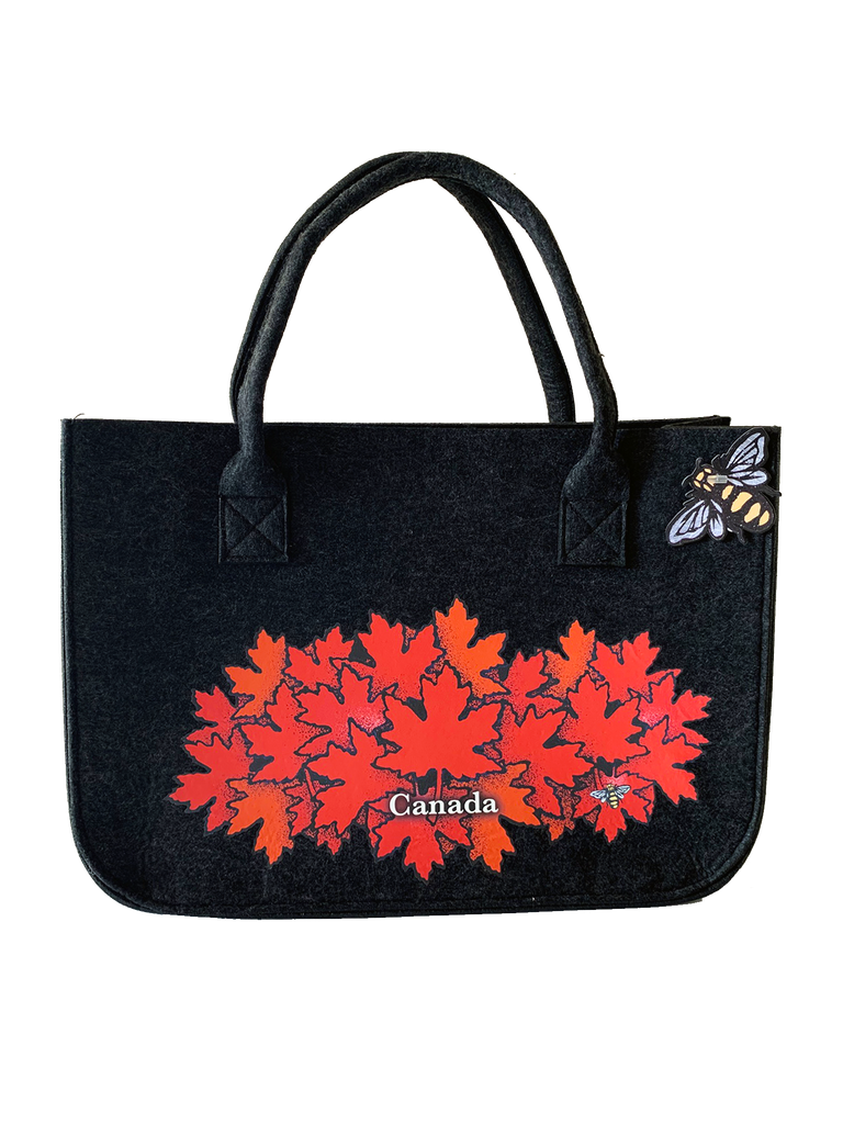 This large dark grey felt bag has two overhead handles and features an art print of a spray of red maple leaves. At the bottom of the leaf pile the word “Canada” has been written in white text. At the bottom right of the picture is the artists mark—a small picture of a bee.