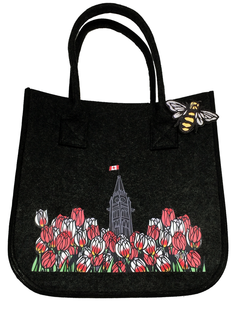 This small dark grey felt bag has two overhead handles and features an art print of the peace tower and a field of tulips. The peace tower is in the center of the picture and is flying the Canadian flag. The dense field of flowers has red tulips and Canada 150 tulips, which are white with fiery streaks of red. At the bottom right of the picture is the artists mark—a small picture of a bee.