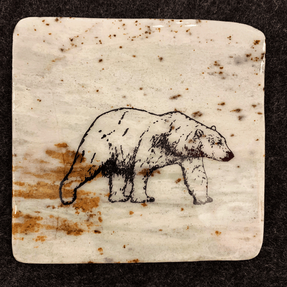This coaster features the image of the side view of a polar bear as it walks. The image is on a piece of canadian shield marble with mineral lines running through in unique colours, lines, and patterns. The coaster is finished with a clear coat, giving it a shiny finish. 