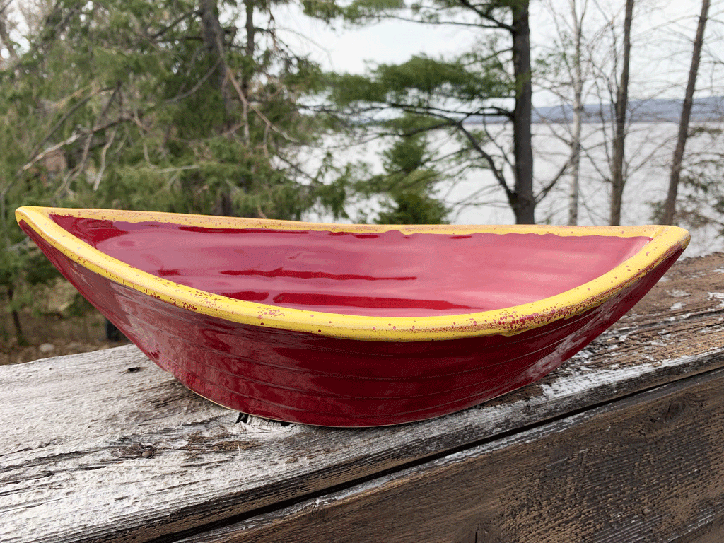 Bowl is red with a yellow rim on the top edges. Bowl is oval shaped with points at two opposite ends. Bowl is on a piece of wood with trees and a lake in the background.