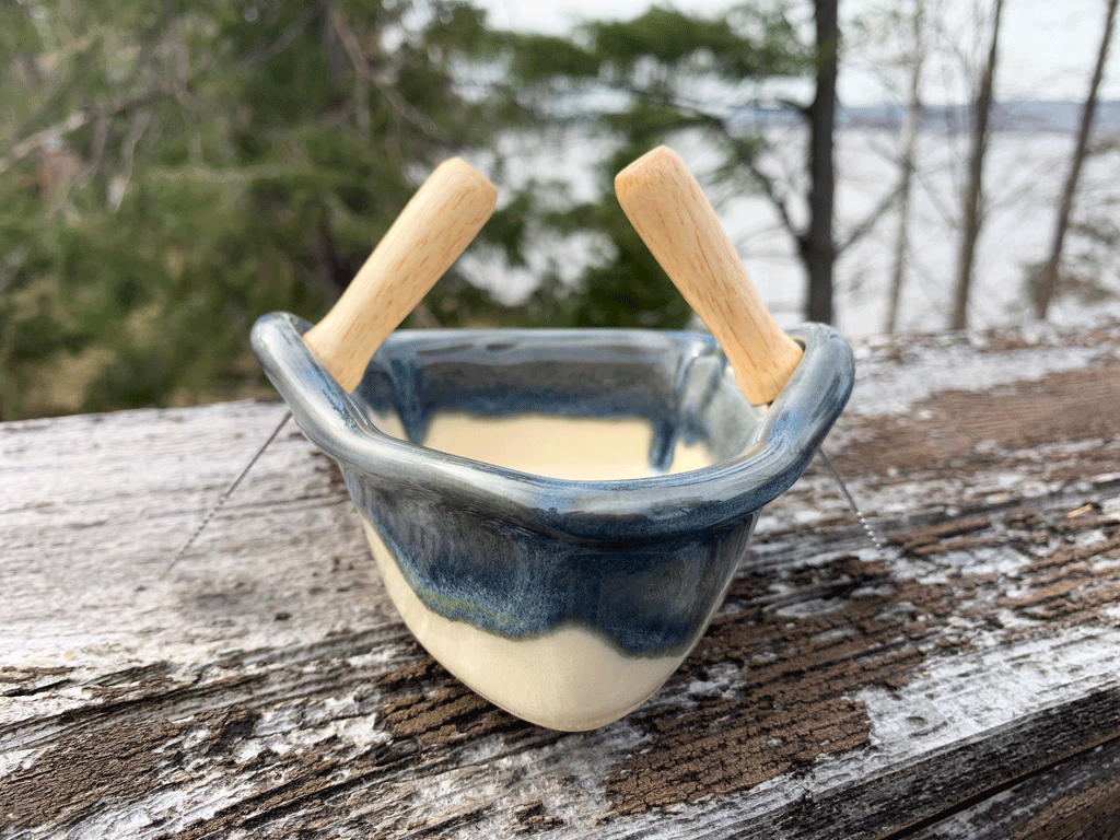 A stoneware rowboat sits on a weathered wooden railing. Two spreaders shaped like paddles sit in the oarlocks. The base is natural white clay, with a contrasting rim in mottled blue and white. A peaceful background of trees and lake complete the picture.