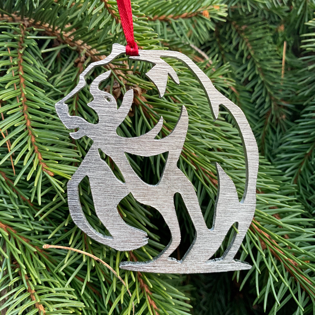 The bear ornament with the ribbon attached and hanging from a pine tree. 