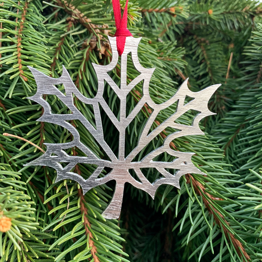The maple leaf ornament with a red ribbon attached and hanging from a pine tree. 