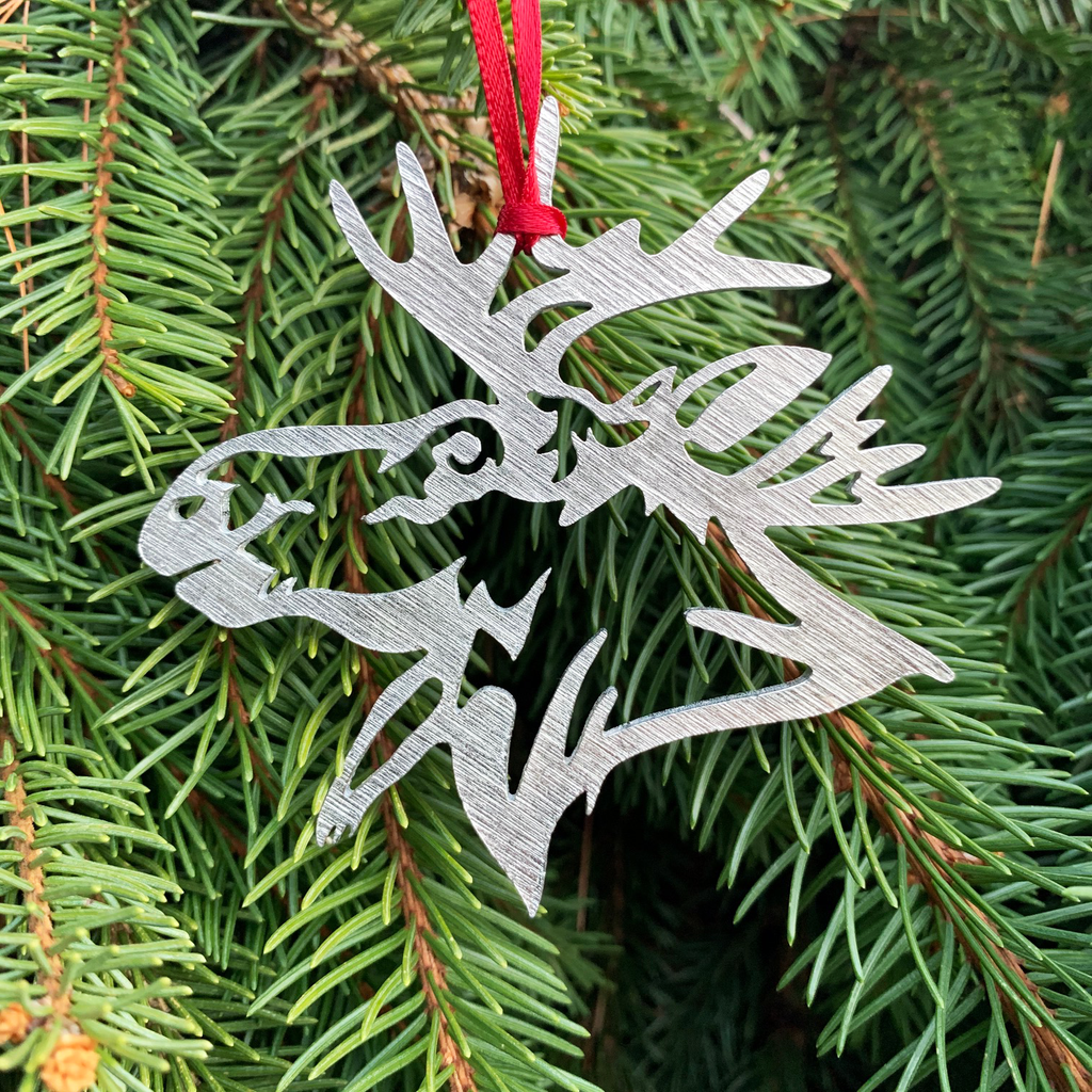 The moose ornament  with a red ribbon attached and hanging from a pine tree. 