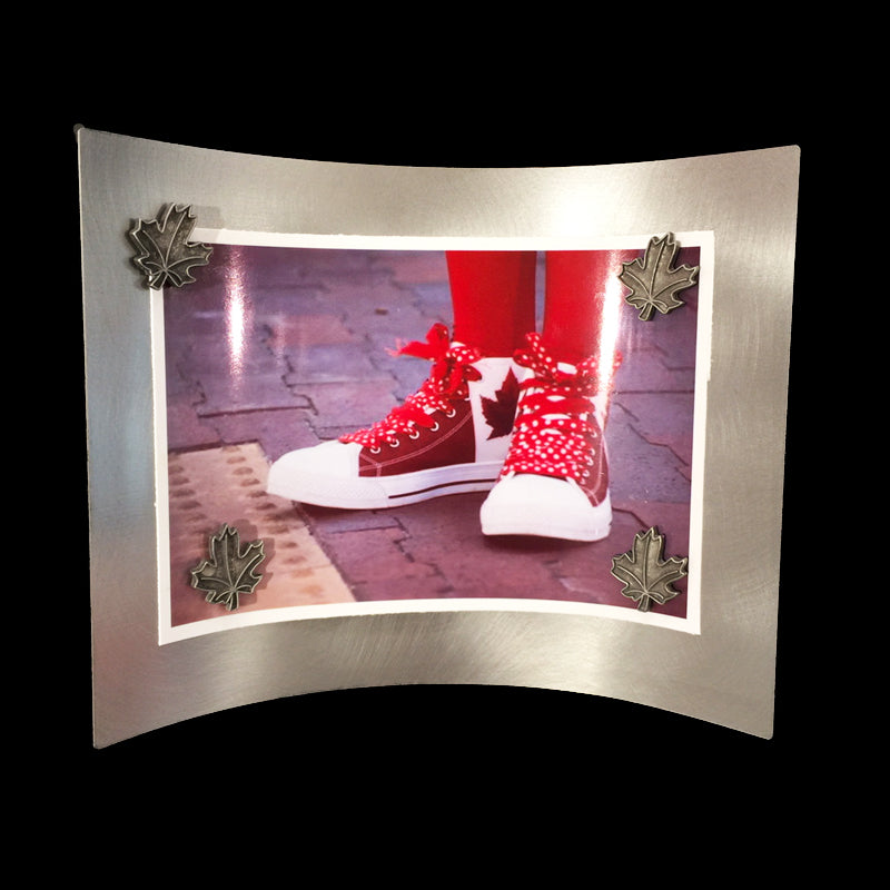 A metal rectangle, curved so it stands without additional pieces supporting it. A photo of someone red and white Canadian flag shoes is held on by four maple leaf shaped pewter magnets. 