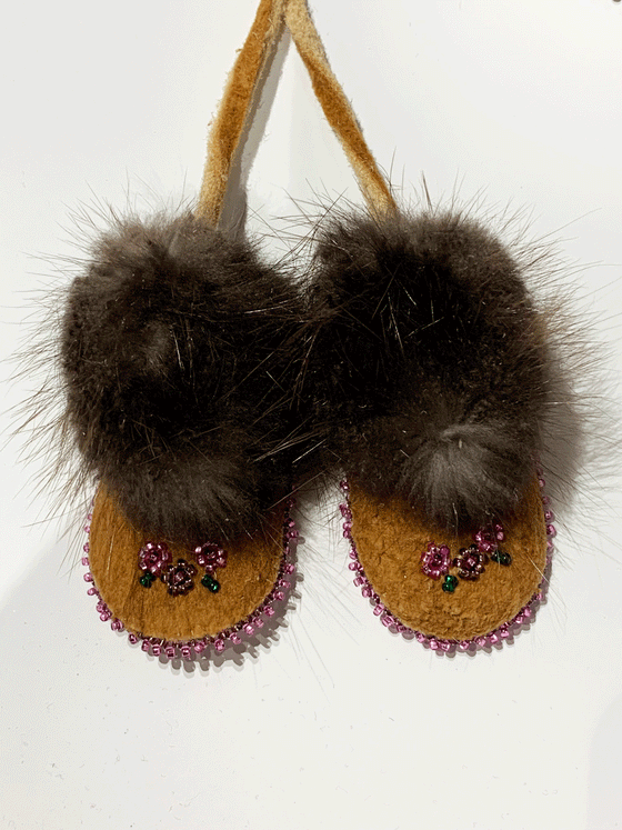 A pair of miniature moccasins hang from a keychain ring by two leather bands. The moccasins have fur-trimmed ankles, and small flowers are embroidered with beads on the top. 