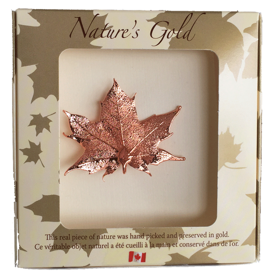 A small copper coated maple leaf sits in a paper box. The top face of the box has a wide square viewing hole. The box is tan with gold maple leaf prints. At the top of the box is written Nature’s Gold. At the bottom is written “this real piece of nature was handpicked and preserved in gold” followed by “Ce véritable objet naturel a été cueilli à la main et conservé dans de l’or”