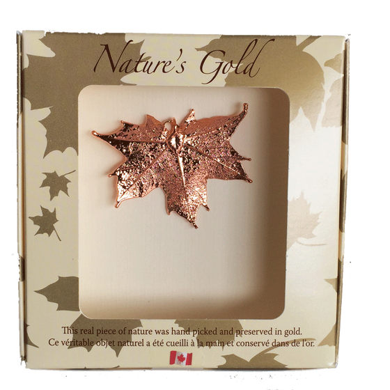 A small copper coated maple leaf sits in a paper box. The top face of the box has a wide square viewing hole. The box is tan with gold maple leaf prints. At the top of the box is written Nature’s Gold. At the bottom is written “this real piece of nature was handpicked and preserved in gold” followed by “Ce véritable objet naturel a été cueilli à la main et conservé dans de l’or”