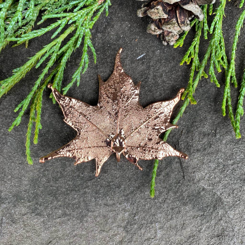 A small copper coated maple leaf sits on a stone background. The copper has a bright finish. At the top of the picture are decorative evergreen leaves and a pine cone.